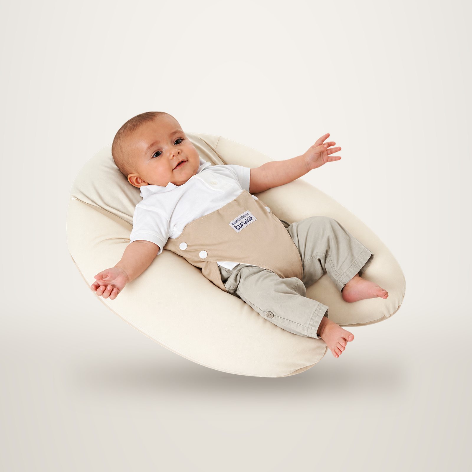 Un bébé en body blanc et pantalon beige est allongé au creux d’un coussin d’allaitement crème, sur fond de studio.