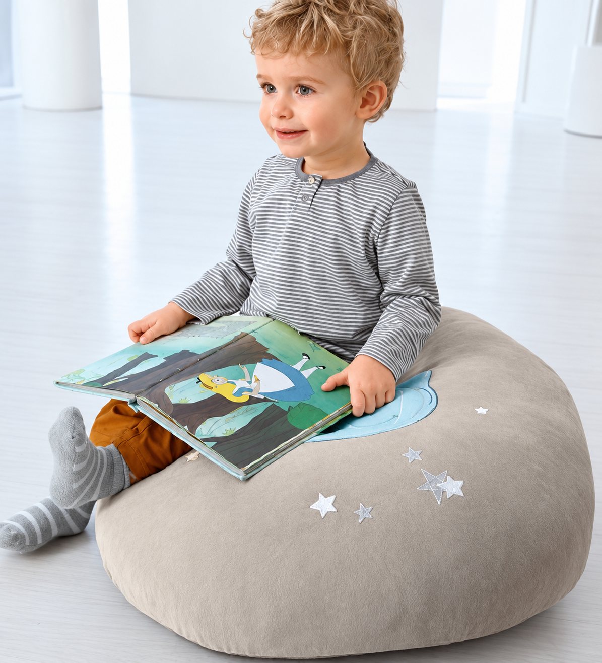 Un petit garçon blond en haut rayé est assis sur un grand pouf beige orné d’étoiles brodées et tient un livre d’images ouvert sur ses genoux.