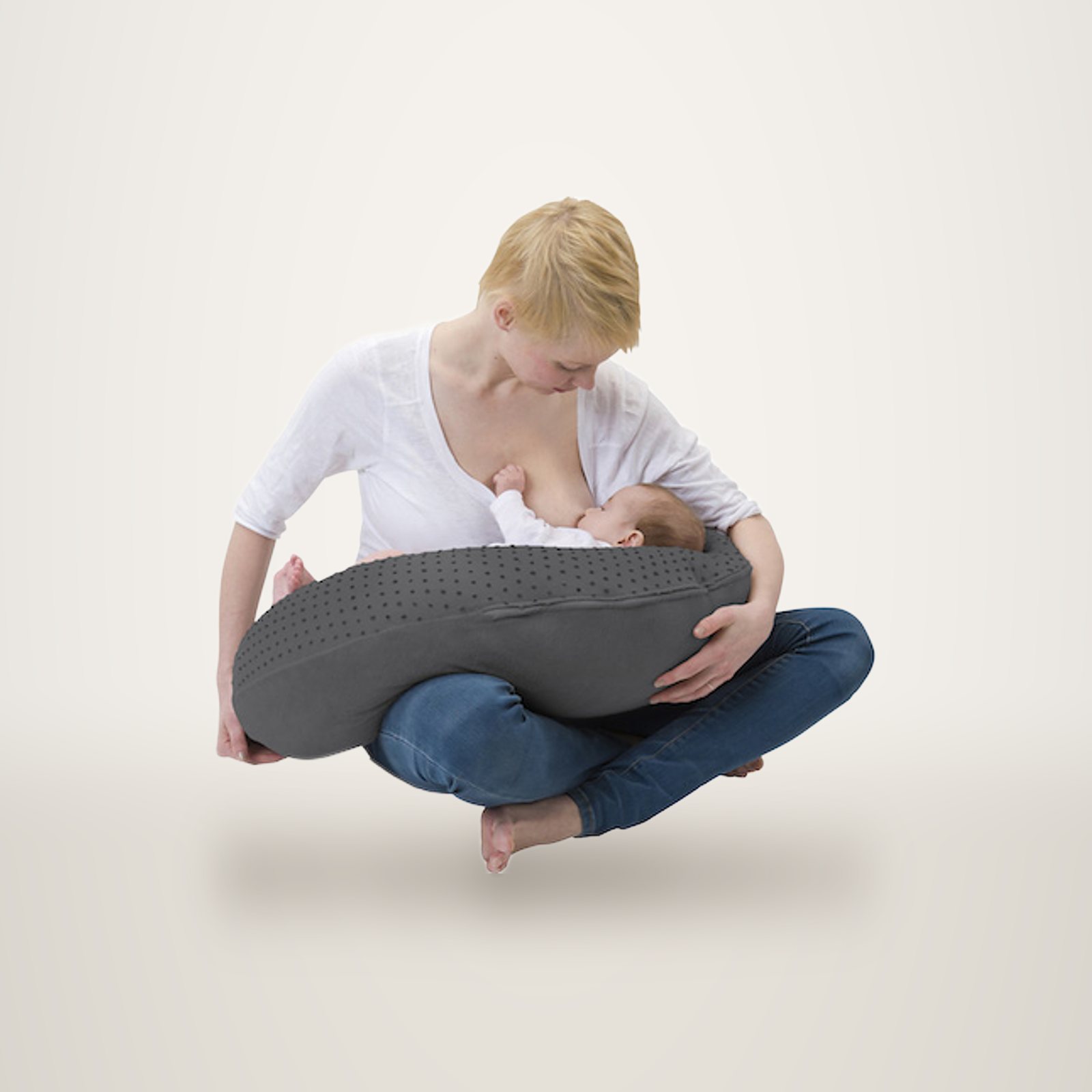 Une jeune femme allaite son bébé sur un grand coussin d’allaitement gris, sur fond de studio.