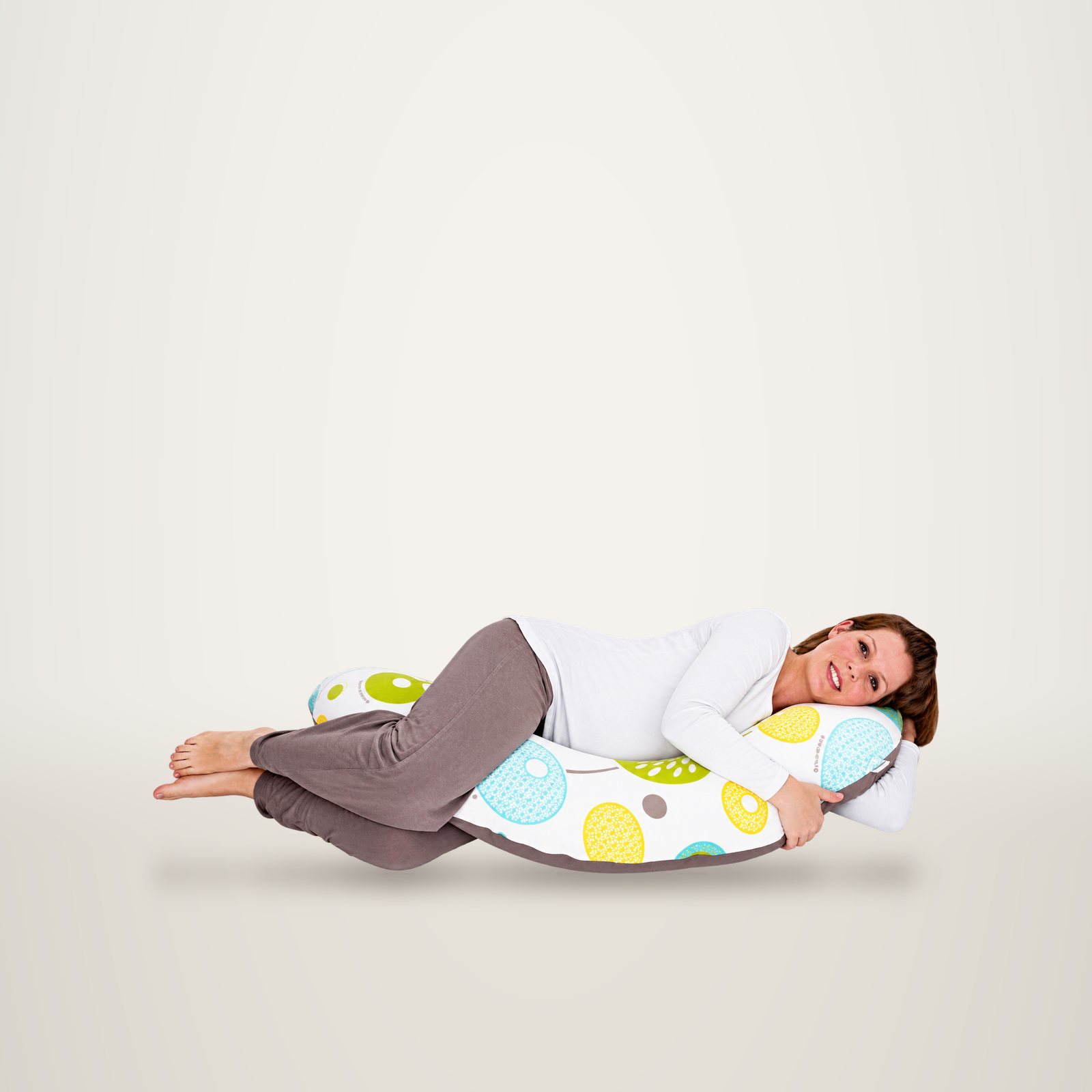 Une femme allongée sur le côté serre un long coussin de maternité blanc à grands motifs ronds colorés, sur fond de studio.