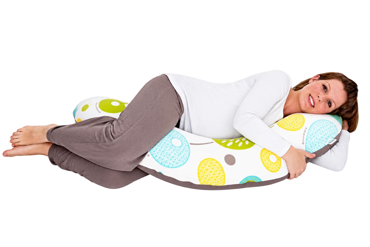 Une femme souriante est allongée sur le côté, serrant un long coussin de maternité blanc orné de grands motifs ronds bleus, jaunes et verts.
