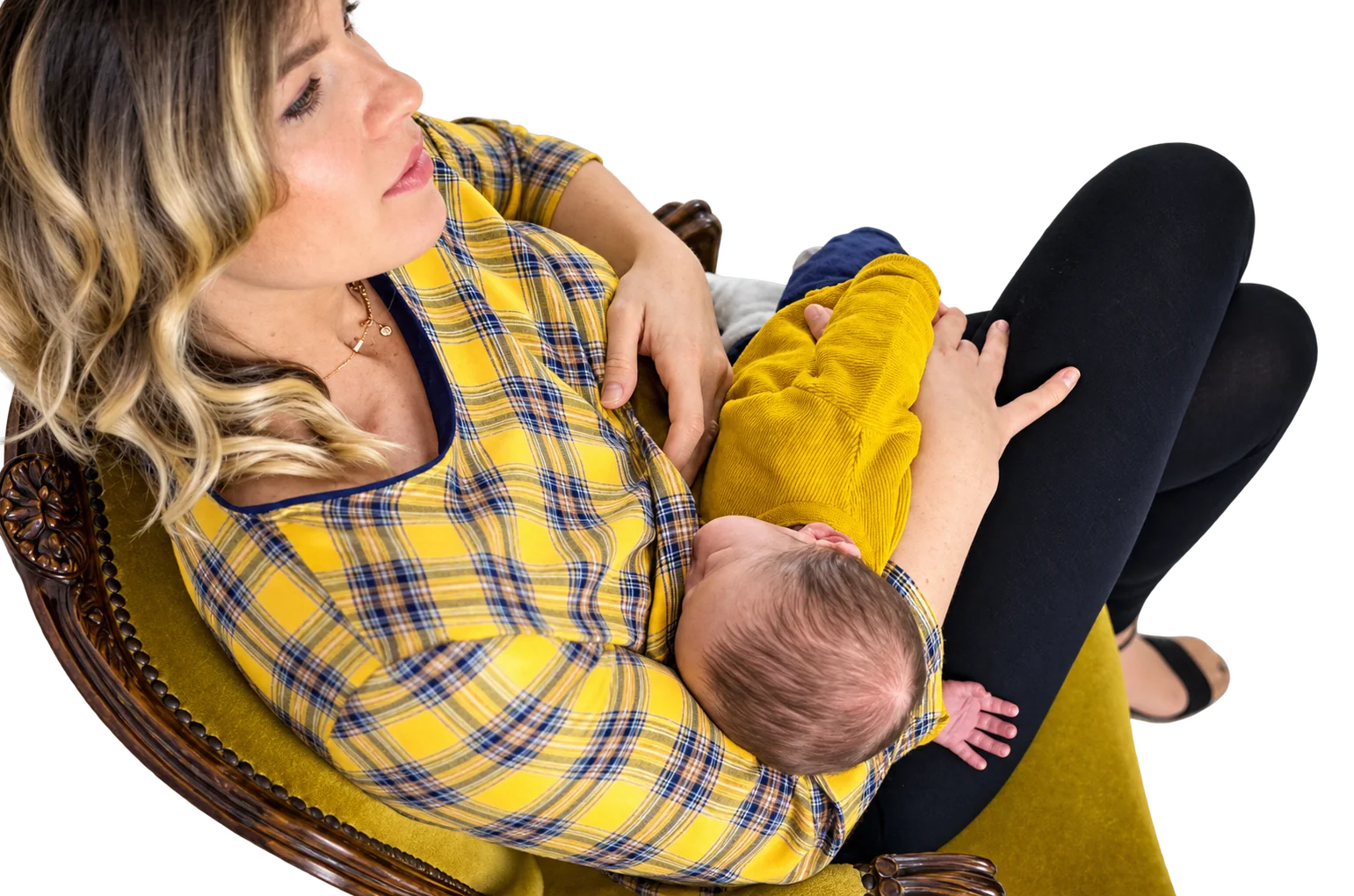 Une femme blonde en chemise à carreaux jaune et bleu tient son nourrisson contre elle, assise dans un fauteuil ancien en velours moutarde.
