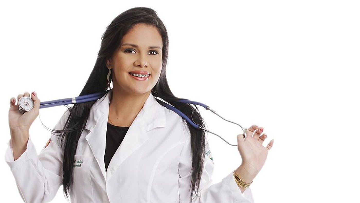 Une femme souriante aux longs cheveux noirs, en blouse blanche, tient un stéthoscope bleu autour de son cou.